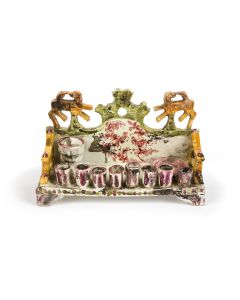 Of sofa form, set on four feet. Painted in multiple colors and glazed (with some color loss.) Row of eight candleholders and featuring miniature goblet and dish set against backplate; flanked by animal (lions?) at top. 5.5 x 8 inches.