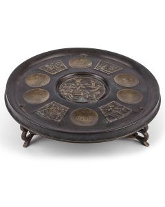 Seder plate with appropriate Passover Seder items labeled in Hebrew and with five Biblical scenes; the whole supported by three-tiered, triangular pedestal to hold matzahs. With Hakishut label on base. Height: 4 inches; Diameter: 12.5 inches.