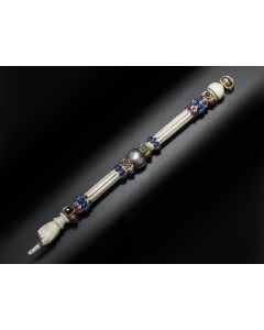 Elaborately decorated with colorful cabochon and faceted gemstones, set along striated shaft with central silver knop with ivory finial; terminating in hand and pointed finger wearing a bejeweled gold ring. Length: 10 inches.